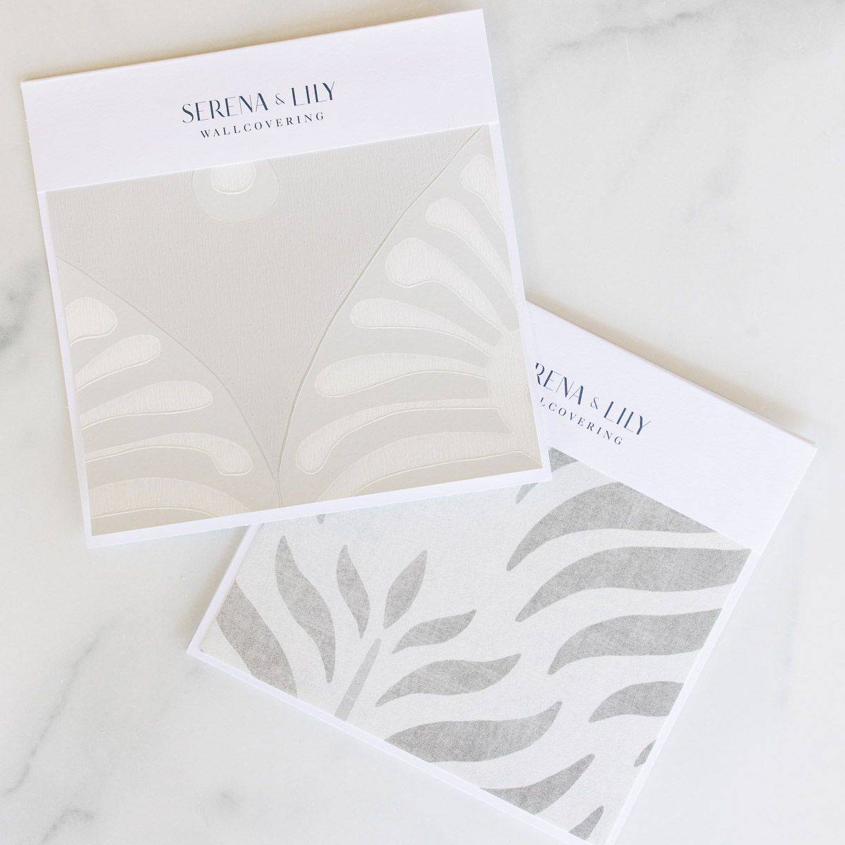 Two neutral patterned Serena and LIly wallpapers laid out on a white marble surface.