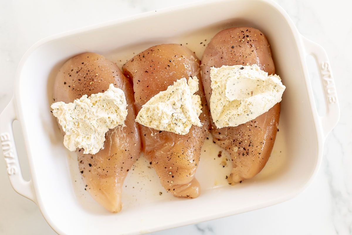 Uncooked chicken breasts in a white baking dish, topped with Boursin cheese.