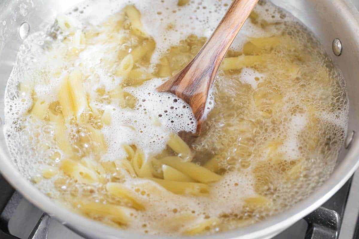 A pot of penne pasta boiling in water with a wooden spoon in the center.