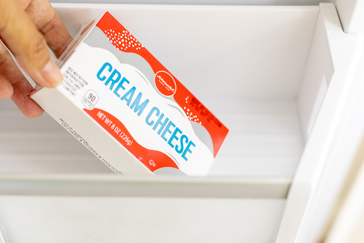 A hand placing a package of cream cheese in a refrigerator drawer to freeze.