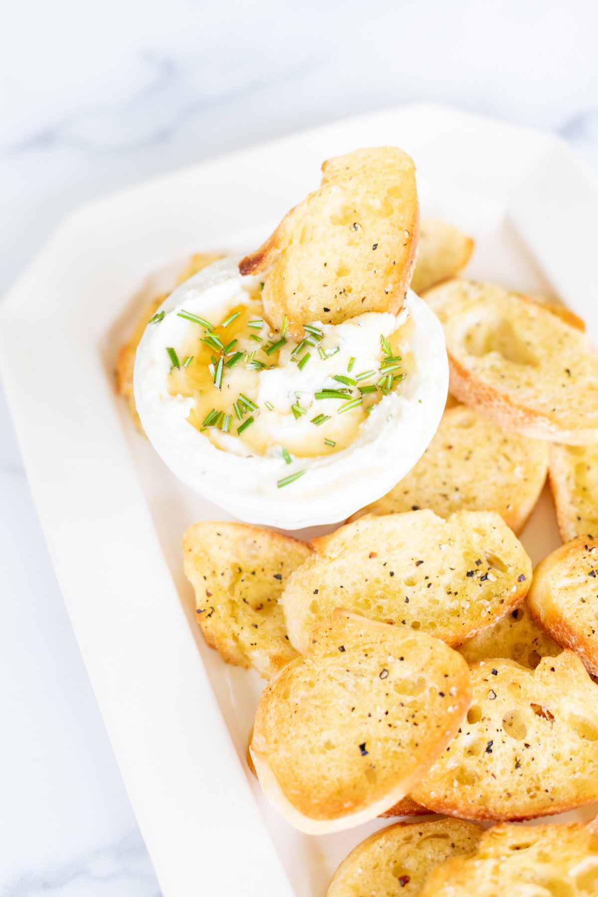 A white dish with a round dip bowl filled with creamy dip topped with chopped herbs and a drizzle of oil, accompanied by multiple toasted crostini on a rectangular white platter.