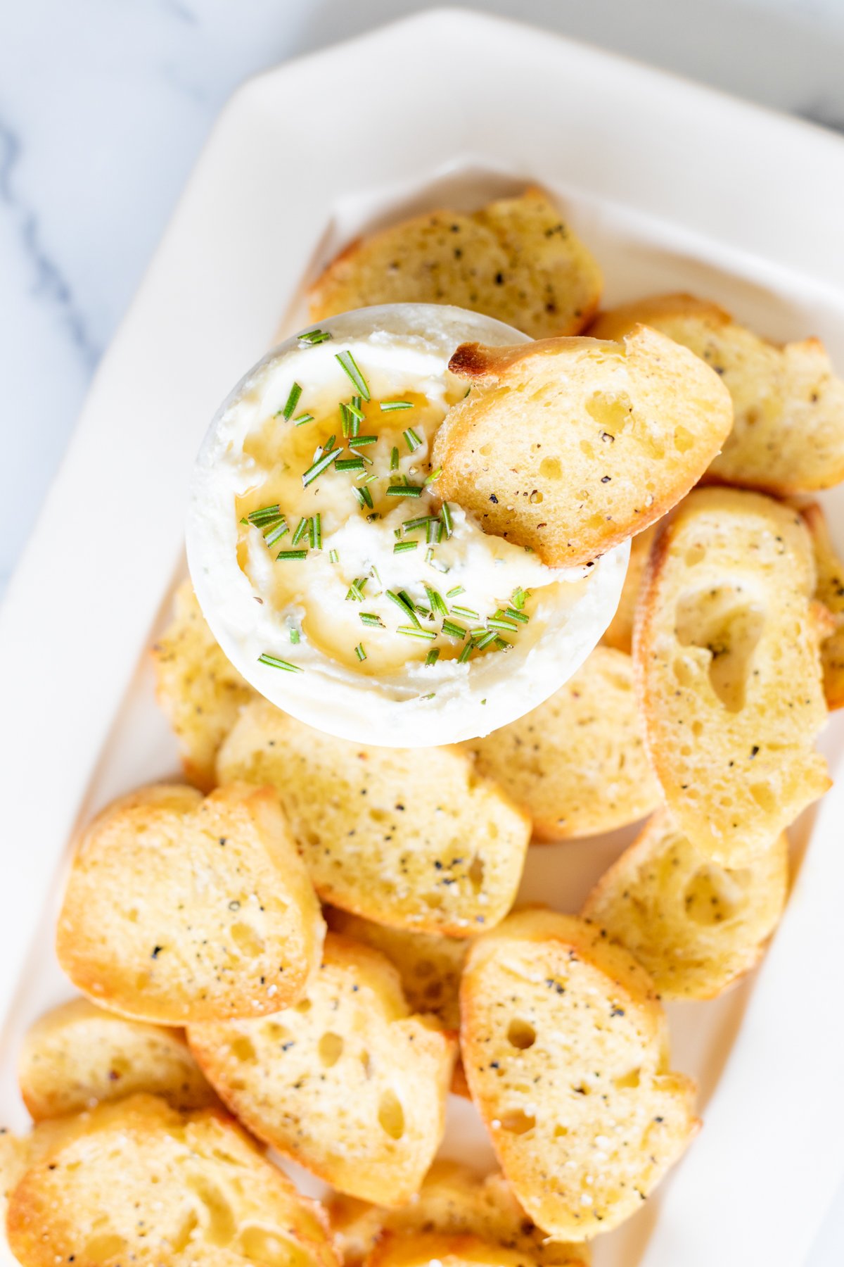 A plate of toasted crostini surrounds a bowl of creamy dip garnished with herbs. One bread slice is dipped into the bowl.