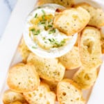 A bowl of creamy dip topped with chopped chives, surrounded by several toasted crostini slices.