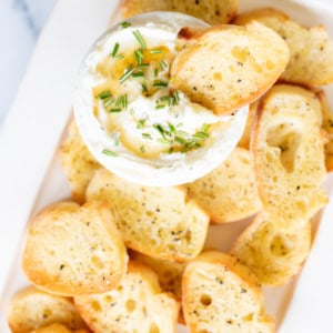 A bowl of creamy dip topped with chopped chives, surrounded by several toasted crostini slices.
