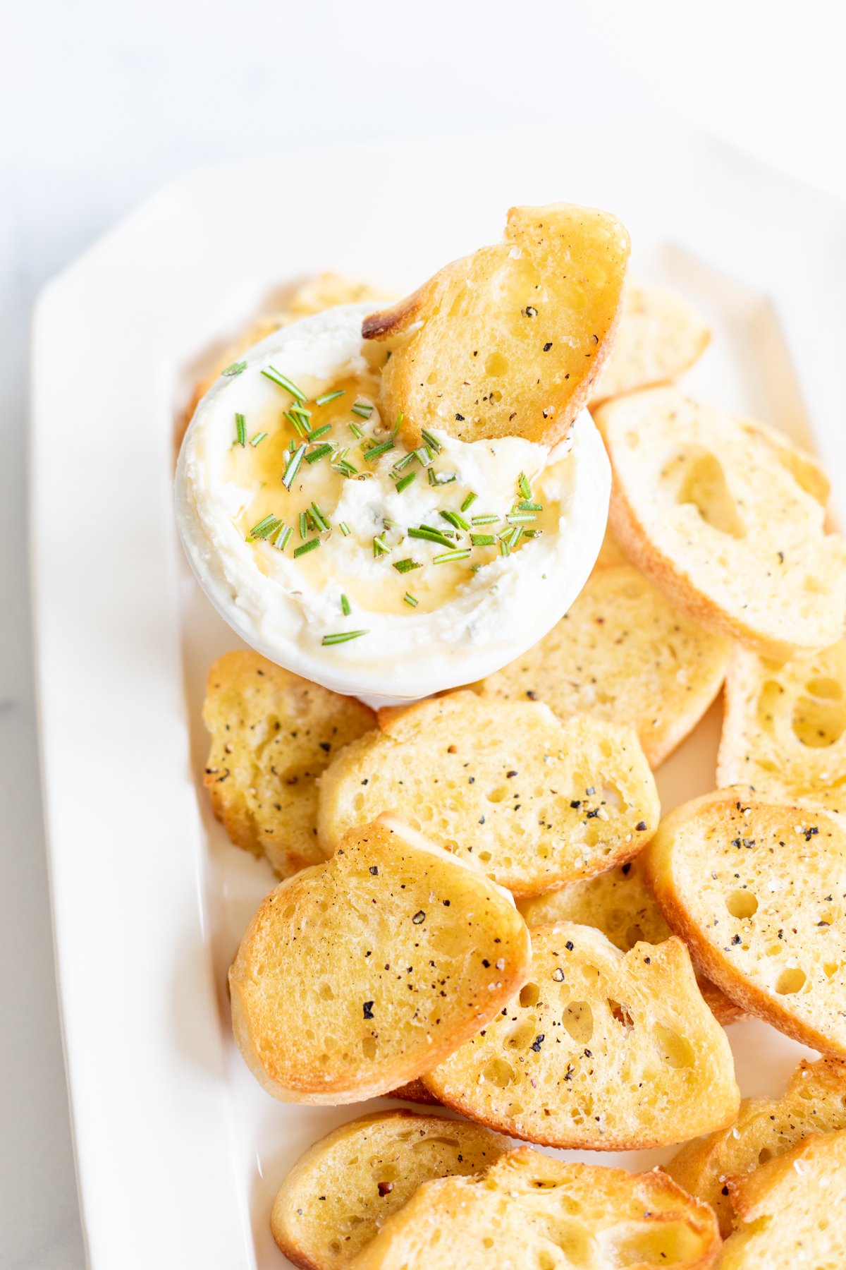 A plate of toasted crostini slices with a small bowl of creamy dip garnished with chopped herbs is displayed.