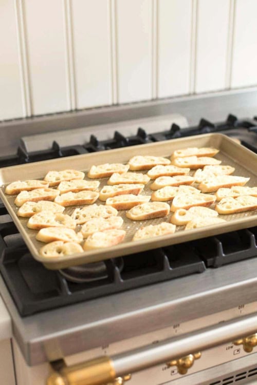 A baking tray with evenly spaced, lightly toasted crostini sits on a gas stove.