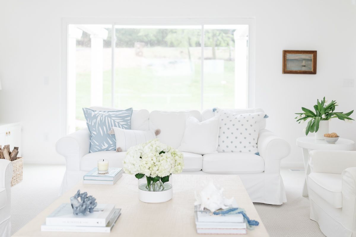 A white living room with a raffia coffee table, with stacks of decorative coffee table books