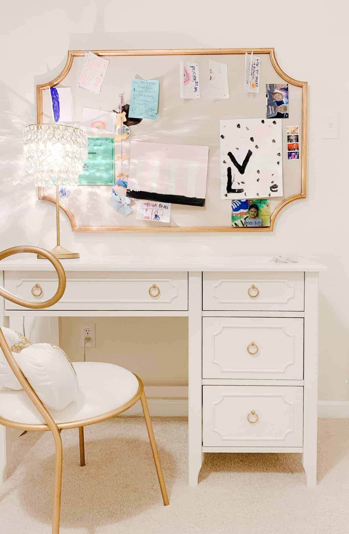 A white desk for teenagers in a girls bedroom, paired with a gold chair and pin board on the wall.