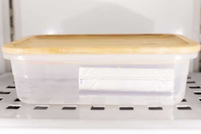 A block of cream cheese wrapped in foil in a plastic container with wooden lid in the freezer.