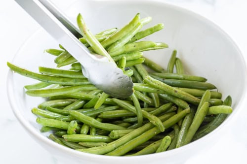 A pair of metal tongs holding grilled green beans over a white bowl filled with more green beans.