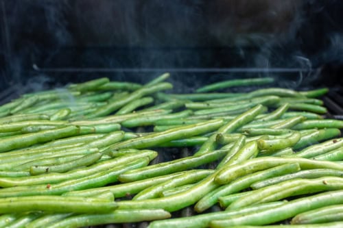 A large number of grilled green beans sizzle on the barbecue, with visible steam rising.