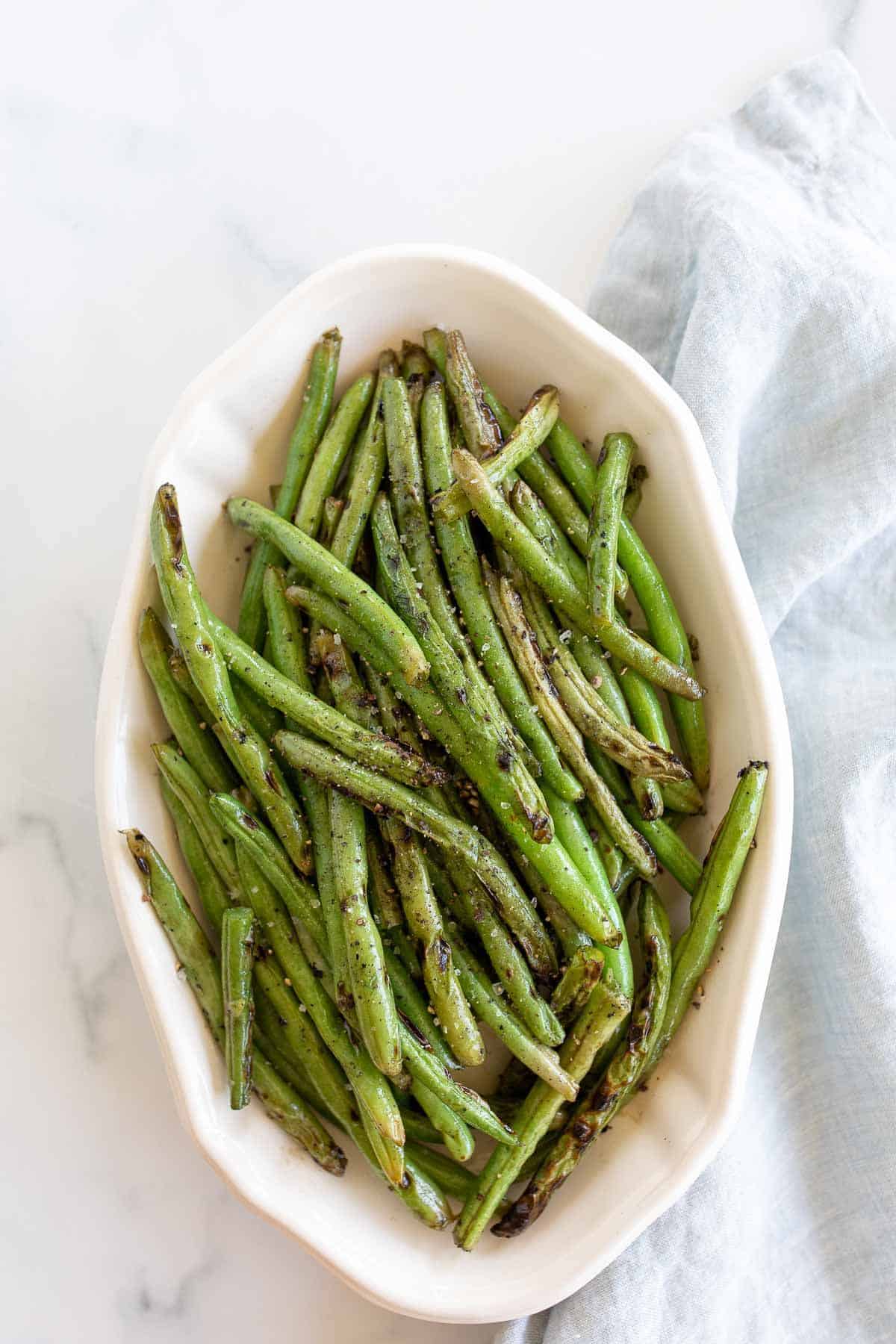 A white oval serving dish, filled with grilled green beans, blue linen towel to the side.