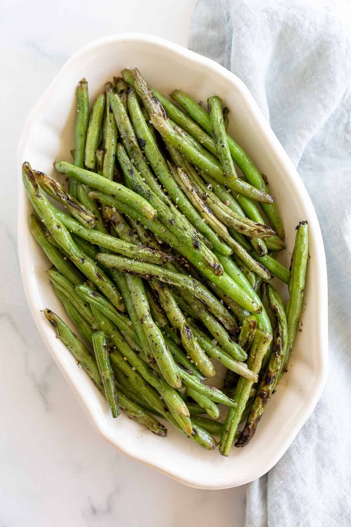 A white oval serving dish, filled with grilled green beans, blue linen towel to the side.