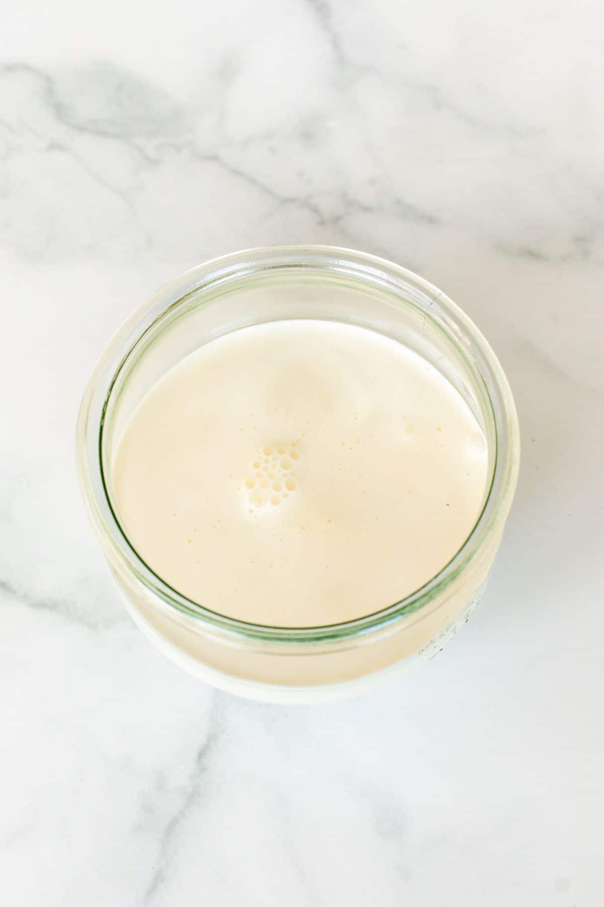 Heavy whipping cream as a creme fraiche substitute in a small clear glass jar on a marble surface.