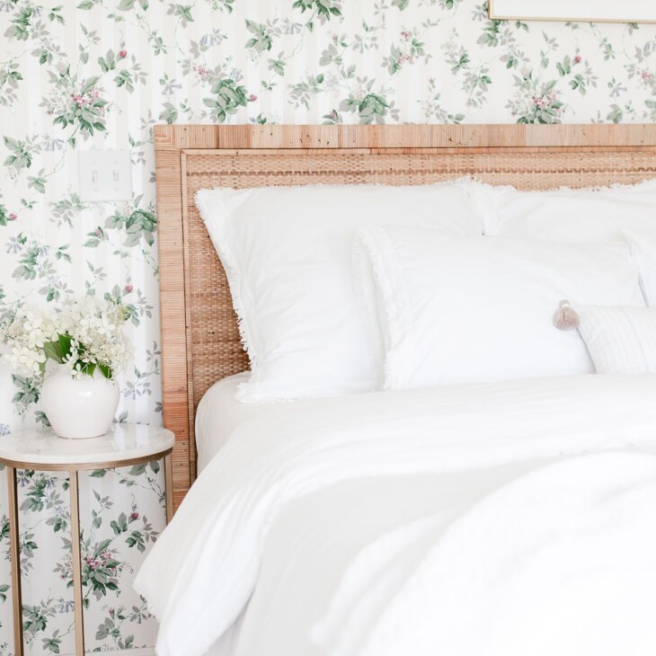 A bedroom with floral wallpaper, a rattan headboard, white bedding and a gold and marble side table.
