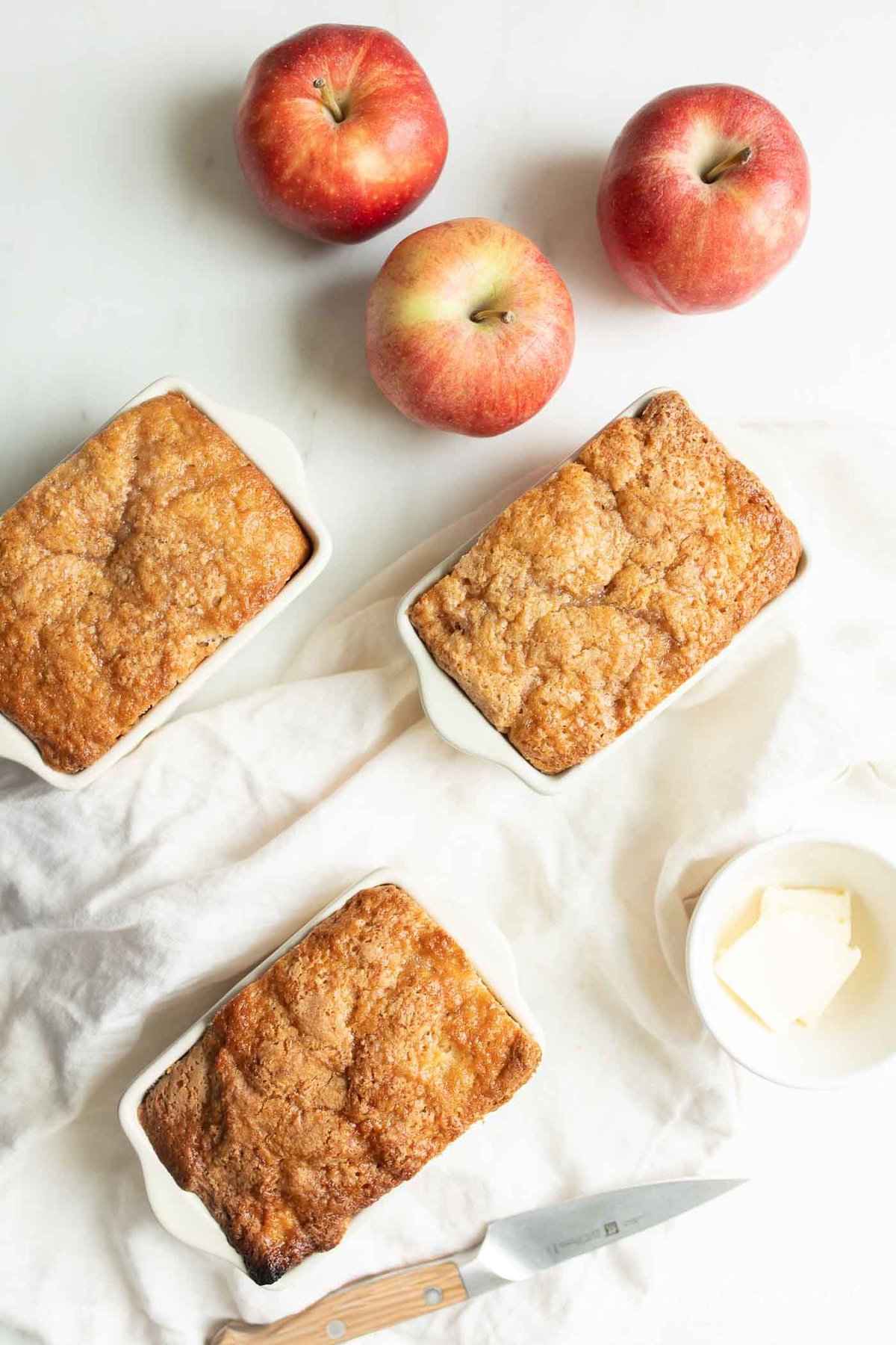 Three small ceramic loaf pans filled with apple bread, with apples in the background