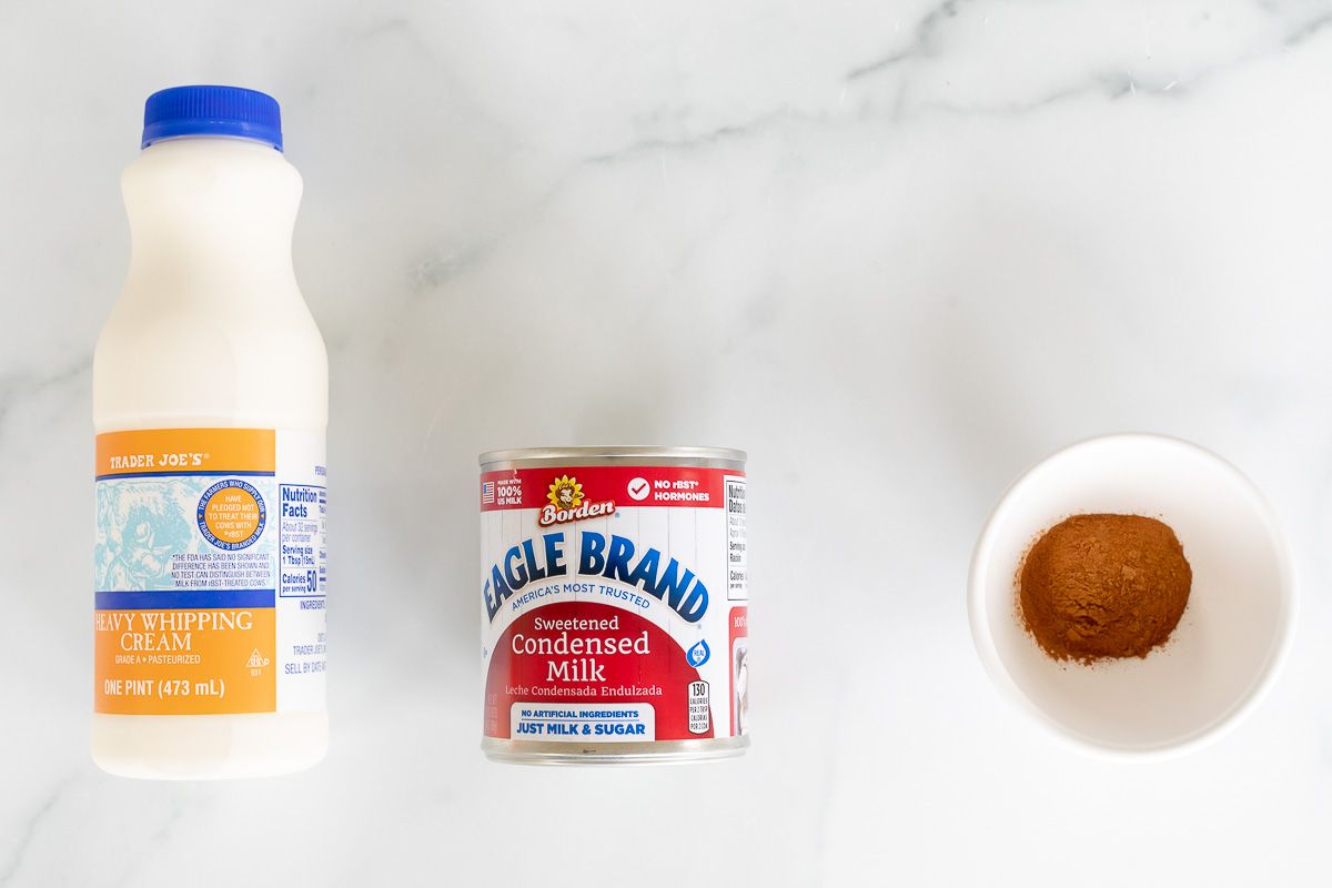 Heavy whipping cream, condensed milk and a bowl of cinnamon laid out on a marble surface.