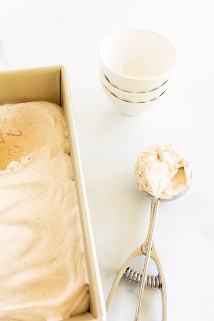 A gold loaf pan full of cinnamon ice cream, with a scoop full to the side, bowls in the background