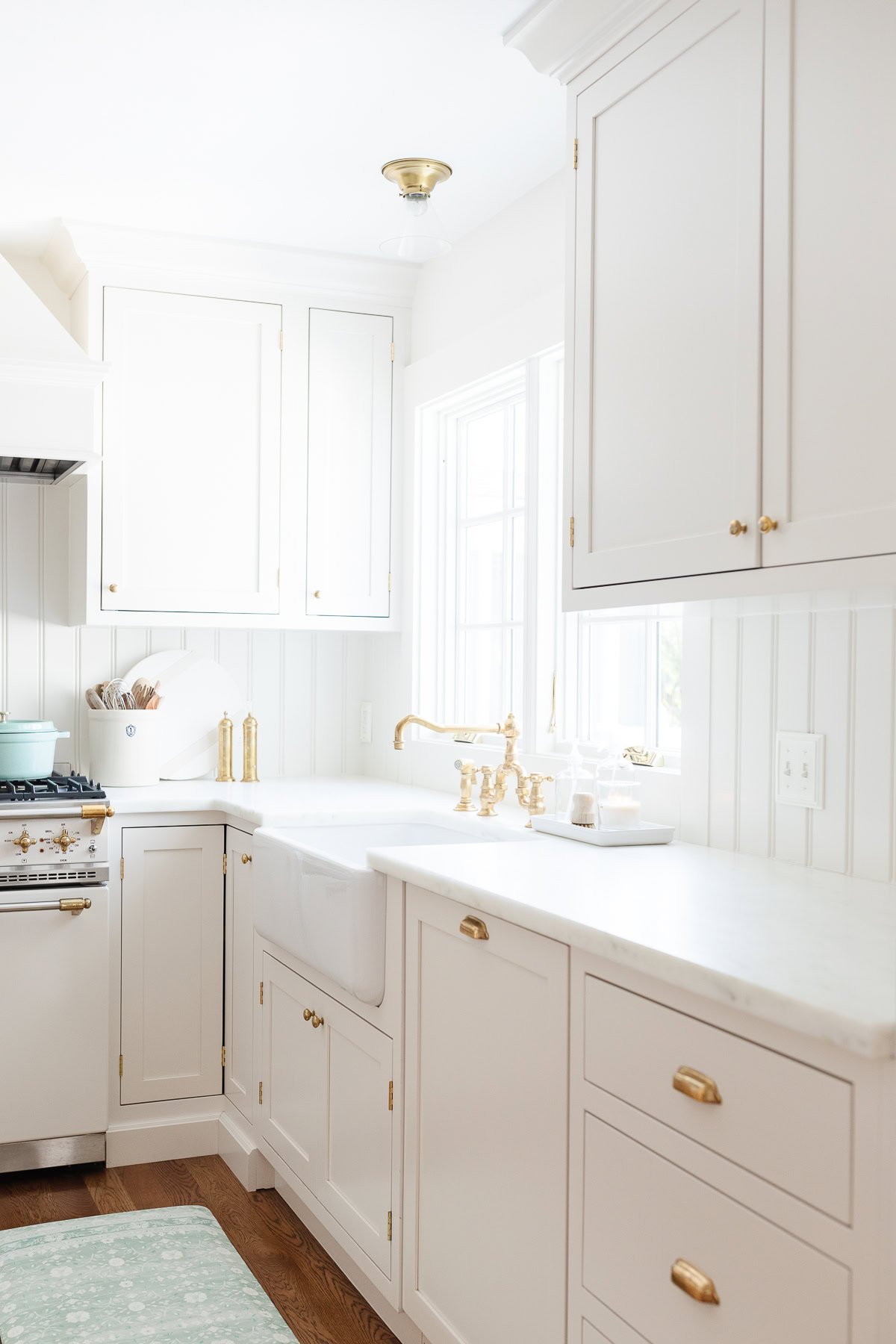 Bright kitchen with white cabinets, gold hardware, farmhouse sink, marble countertop with elegant countertop edges, and a window letting in natural light. Mint green rug and kitchen accessories on the counter.