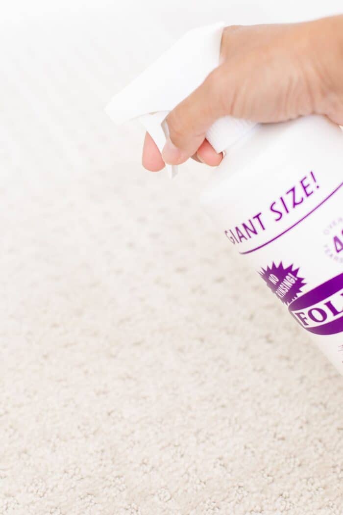 A hand spraying a purple and white bottle of Folex onto a beige patterned carpet.