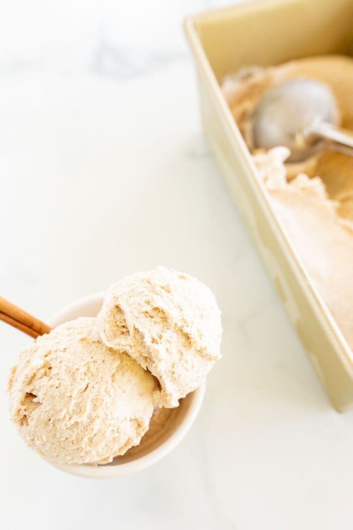 A white bowl full of homemade cinnamon ice cream with a cinnamon stick as garnish on the side, a gold loaf pan full of ice cream in the background