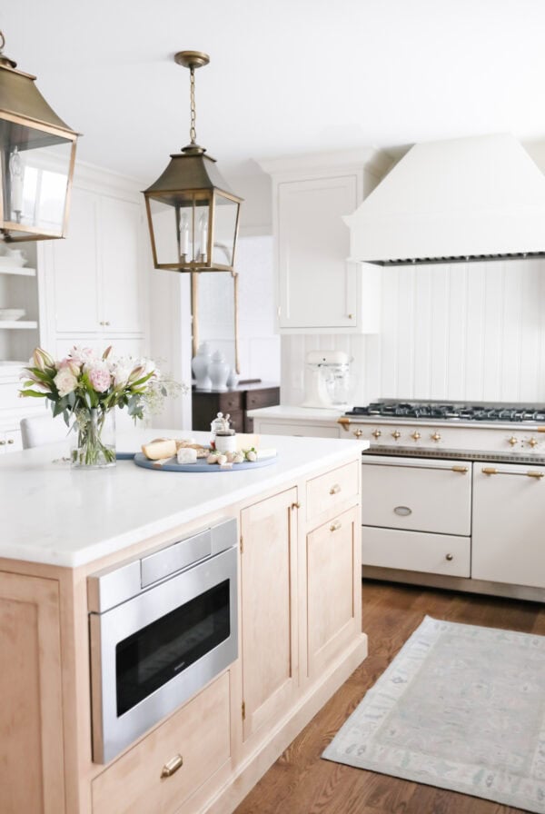 Bright kitchen with white cabinets, a center island, brass light fixtures, a drawer microwave, gas range stove, and a tray with flowers and snacks on the countertop.