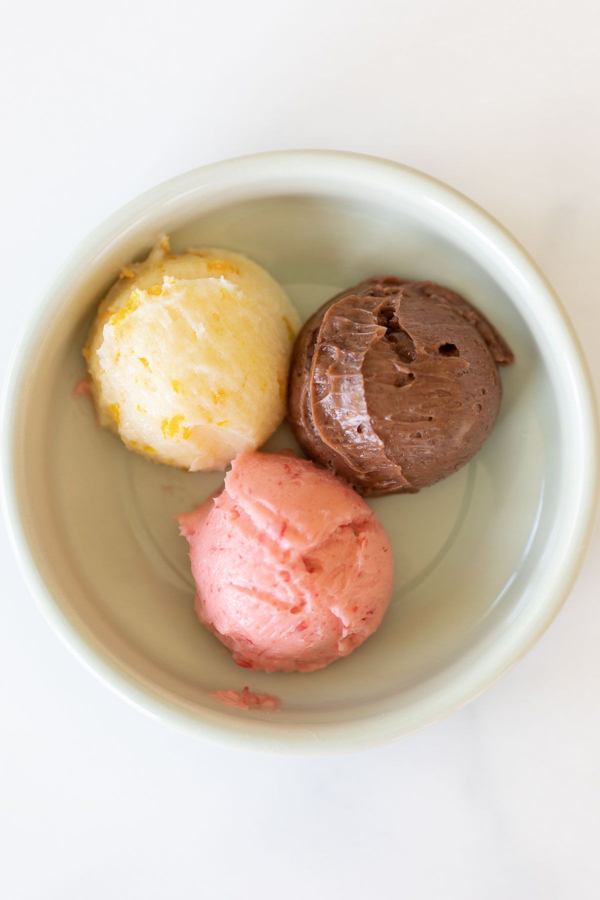 A white bowl on a marble surface, filled with three scoops of flavored butters.