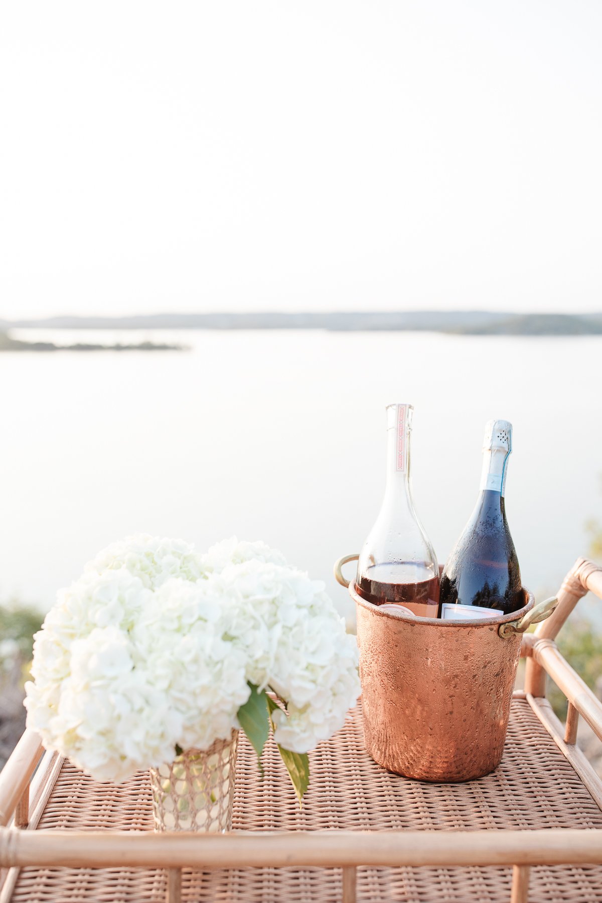 A copper bucket of champagne near a vase of white hydrangeas on a rattan bar cart, water views in the background