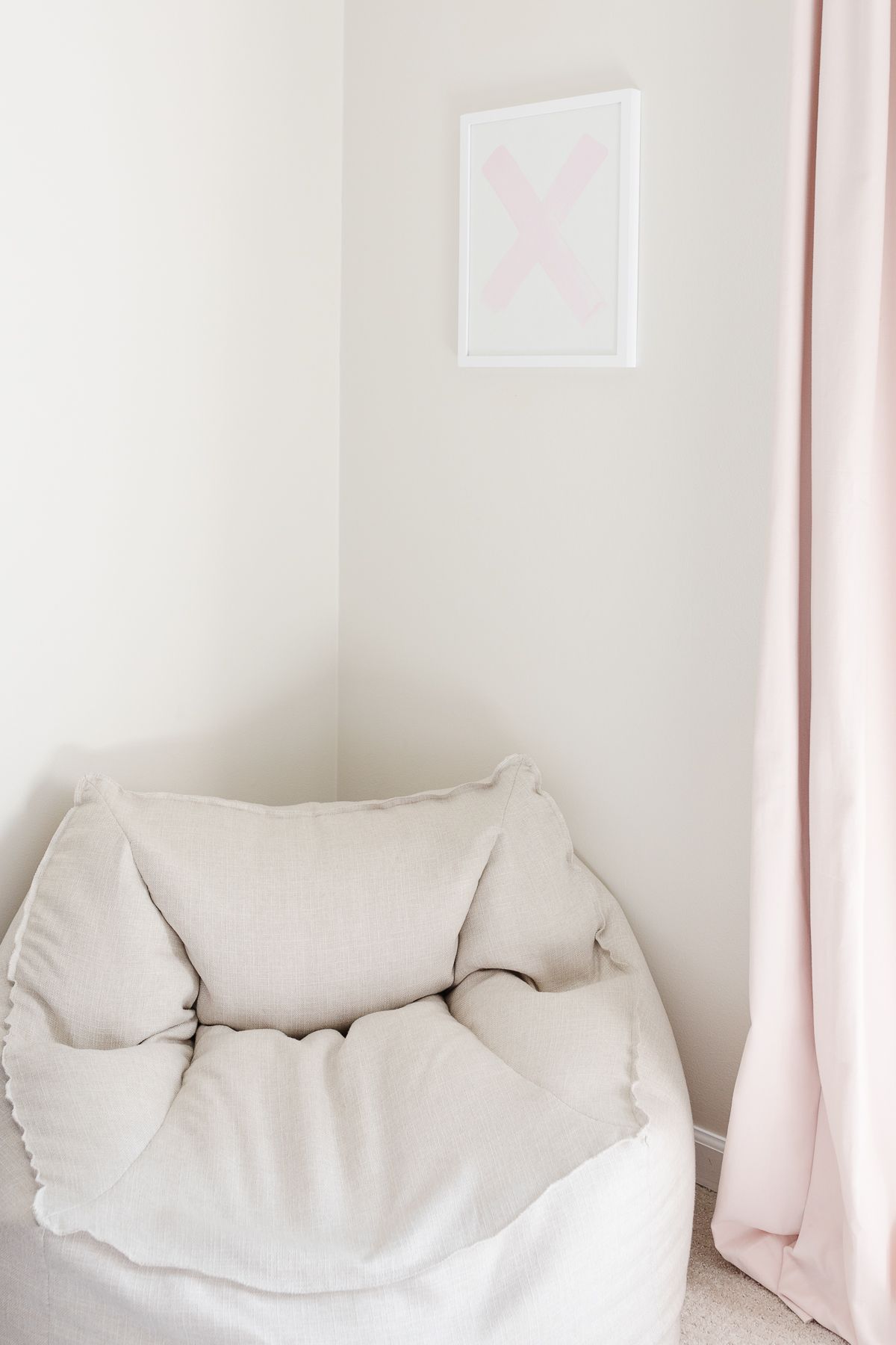 A neutral bean bag chair in the corner of a cream tween bedroom, pink curtain panel on the edge of image.