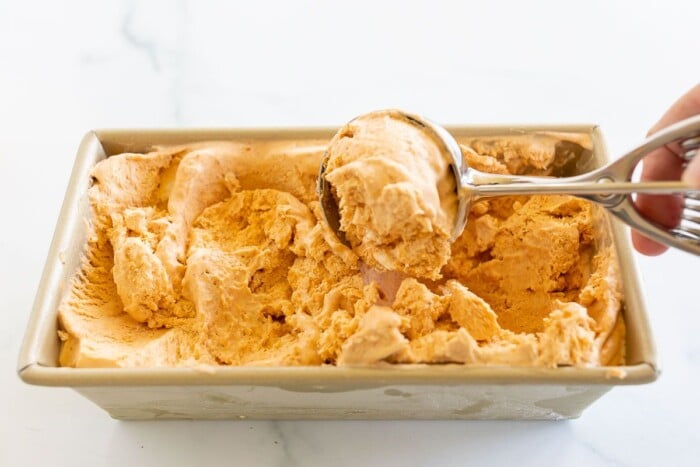 A gold loaf pan filled with pumpkin ice cream, scooper pulling a scoop out.