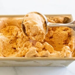A gold loaf pan filled with pumpkin ice cream, scooper pulling a scoop out.