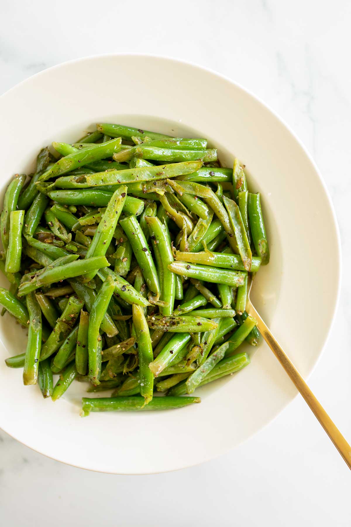 Seasoned green beans in a white bowl with a gold spoon.