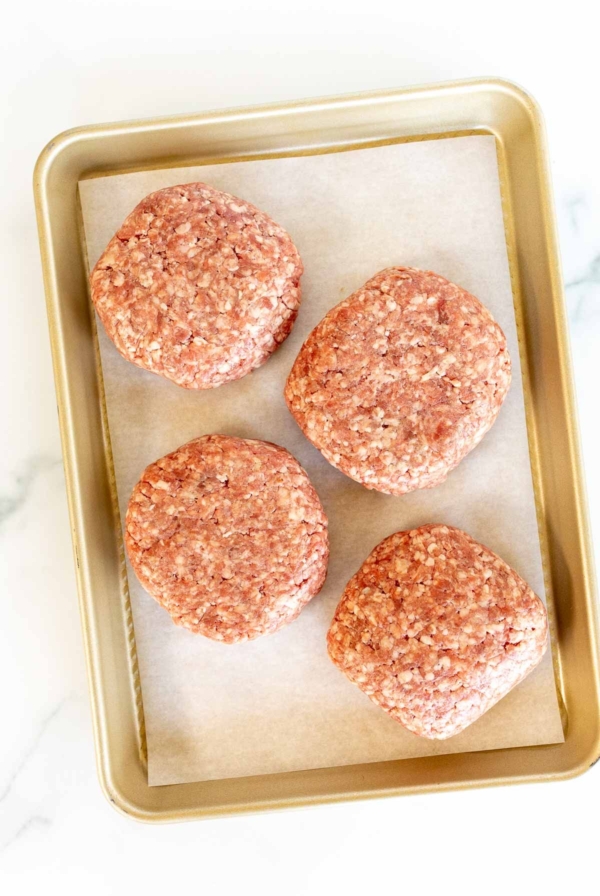 A gold baking sheet with four burger patties on parchment paper.