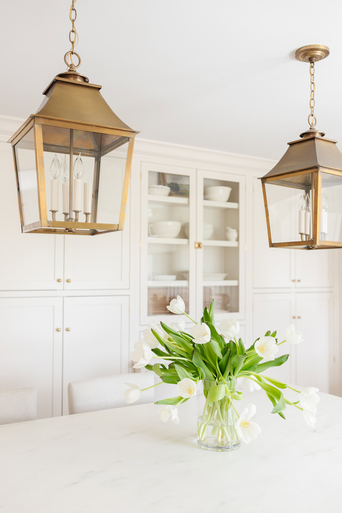 A glass vase of white tulips sits on a marble kitchen island beneath two brass pendant lights, with white cabinets and dishware in the background—perfect inspiration for learning how to measure furniture for your own elegant space.