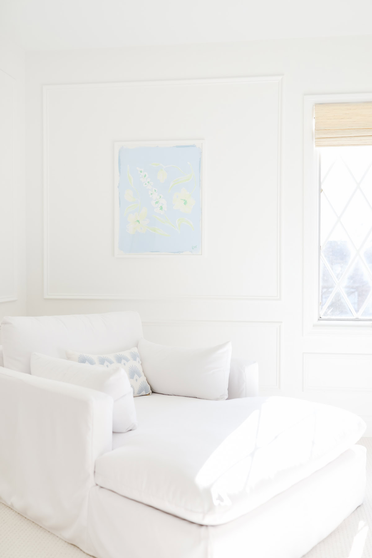 A white armchair with a patterned pillow sits in a bright, white room. If you’re wondering how to measure furniture for your space, notice the compact fit beneath a light blue and green painting, beside a window with a bamboo shade.
