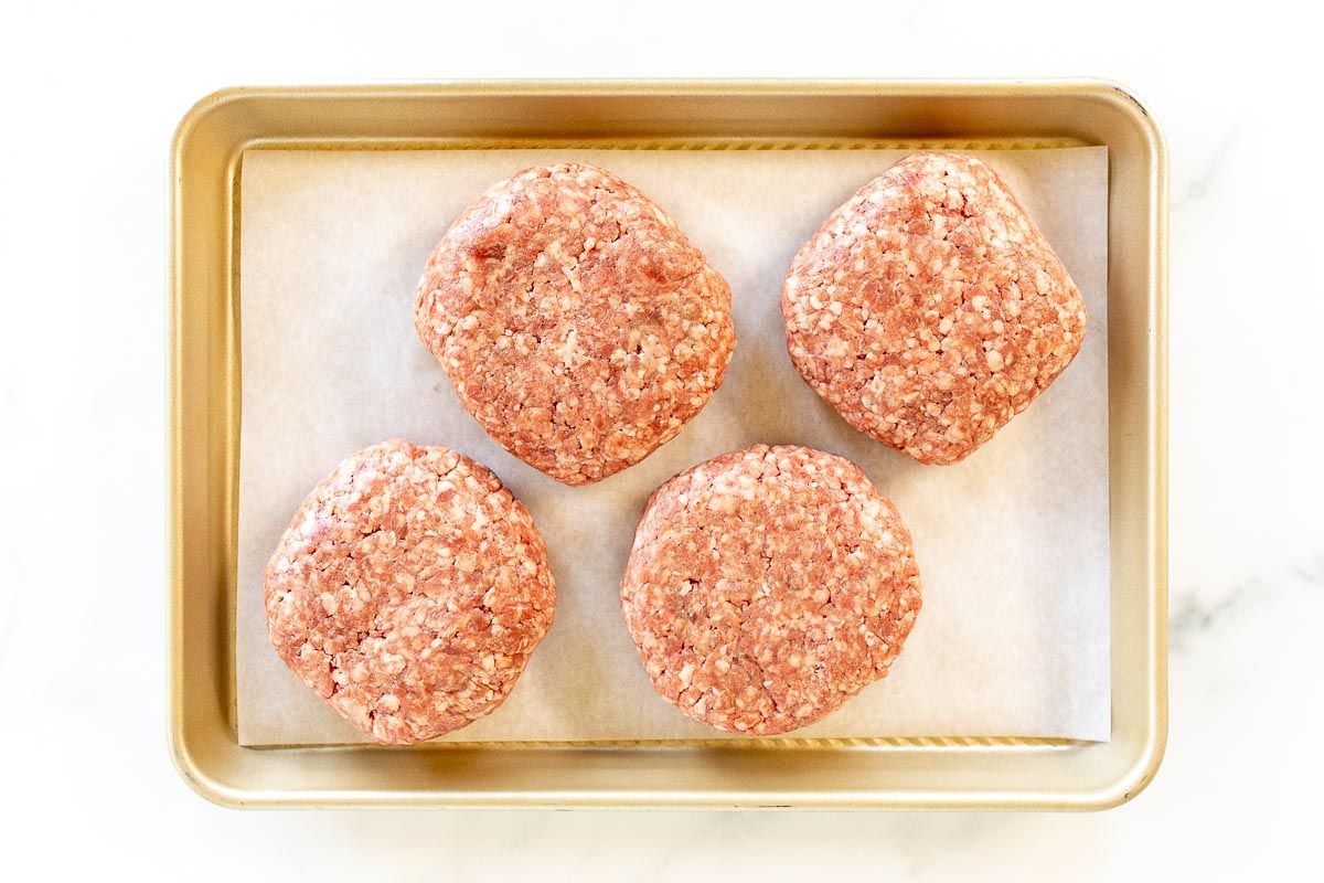A gold baking sheet with four burger patties on parchment paper.