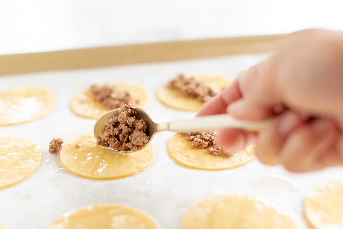 A hand adding ground beef to mini tacos on a parchment lined baking sheet.