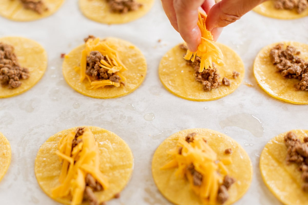 A hand adding cheese to mini tacos on a parchment lined baking sheet.