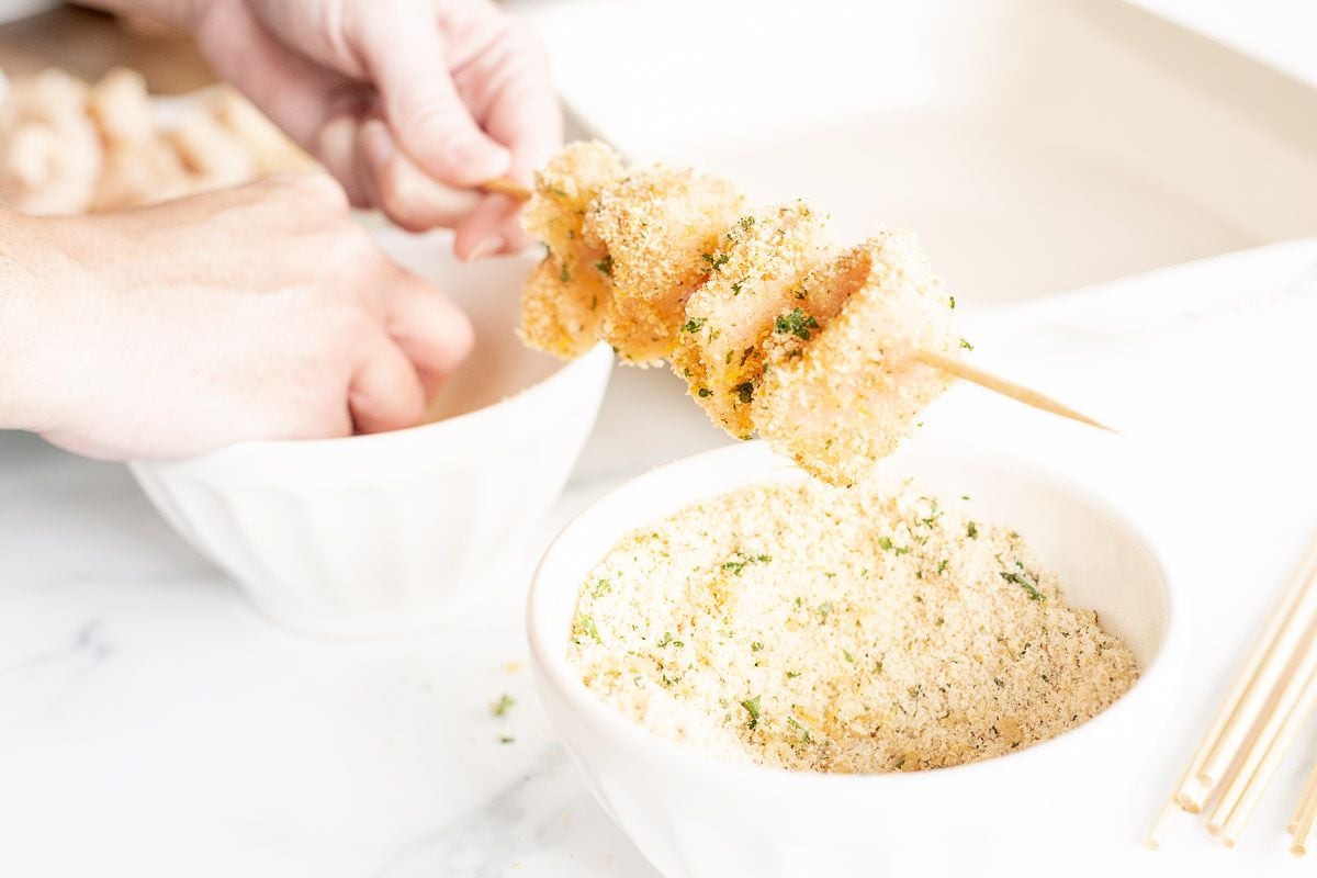 A hand holding a chicken spiedini skewer over a bowl of breading.