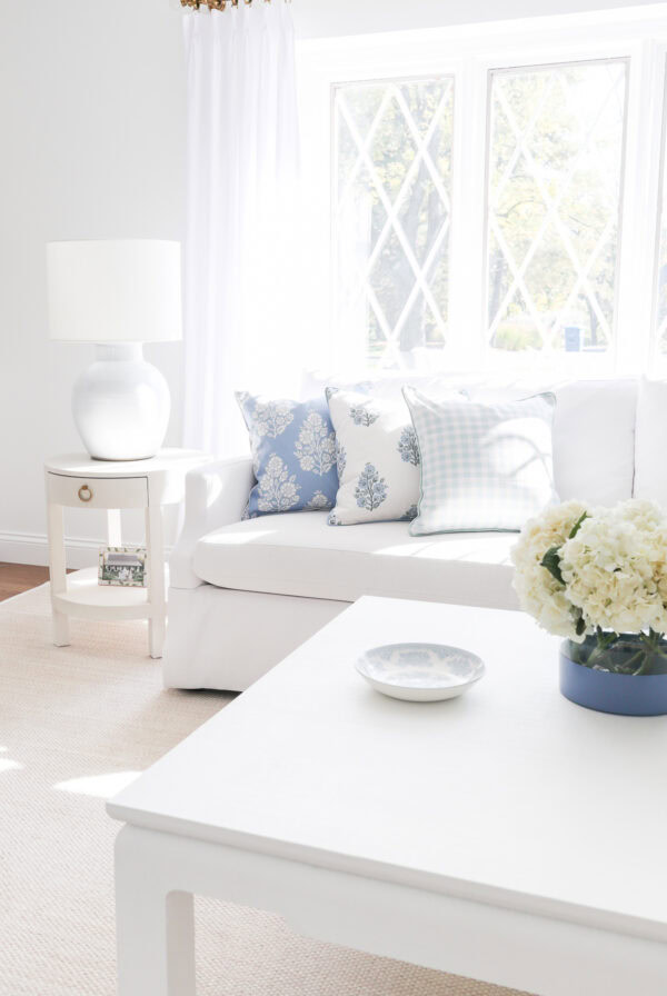 Bright living room with a white sofa, blue and white pillows, a coffee table, and a decorative plate near a window with white curtainsโperfect inspiration if youโre learning how to measure furniture for your own space.