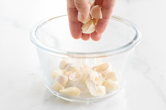 A hand holding garlic cloves, with more cloves in a clear glass bowl.