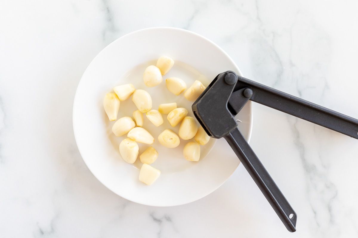 A black garlic press over a white plate full of garlic cloves