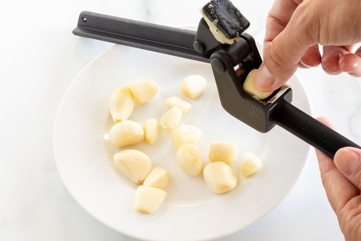 A hand holding a garlic press over a white plate full of garlic cloves.