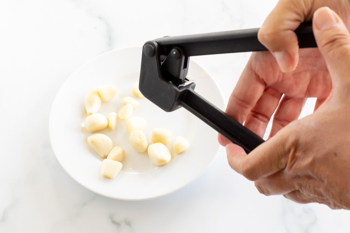 A hand holding a garlic press over a white plate full of garlic cloves.