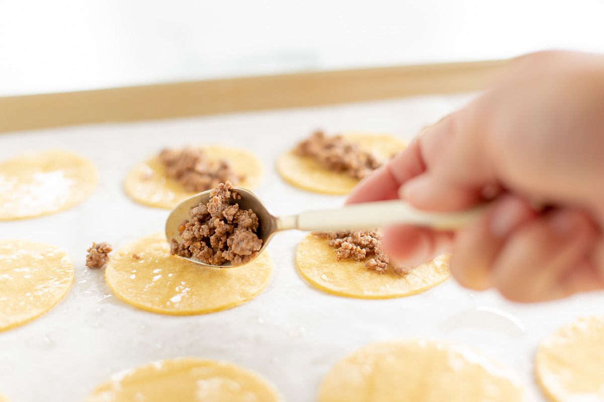 A hand scooping ground beef into mini taco shells on a baking sheet