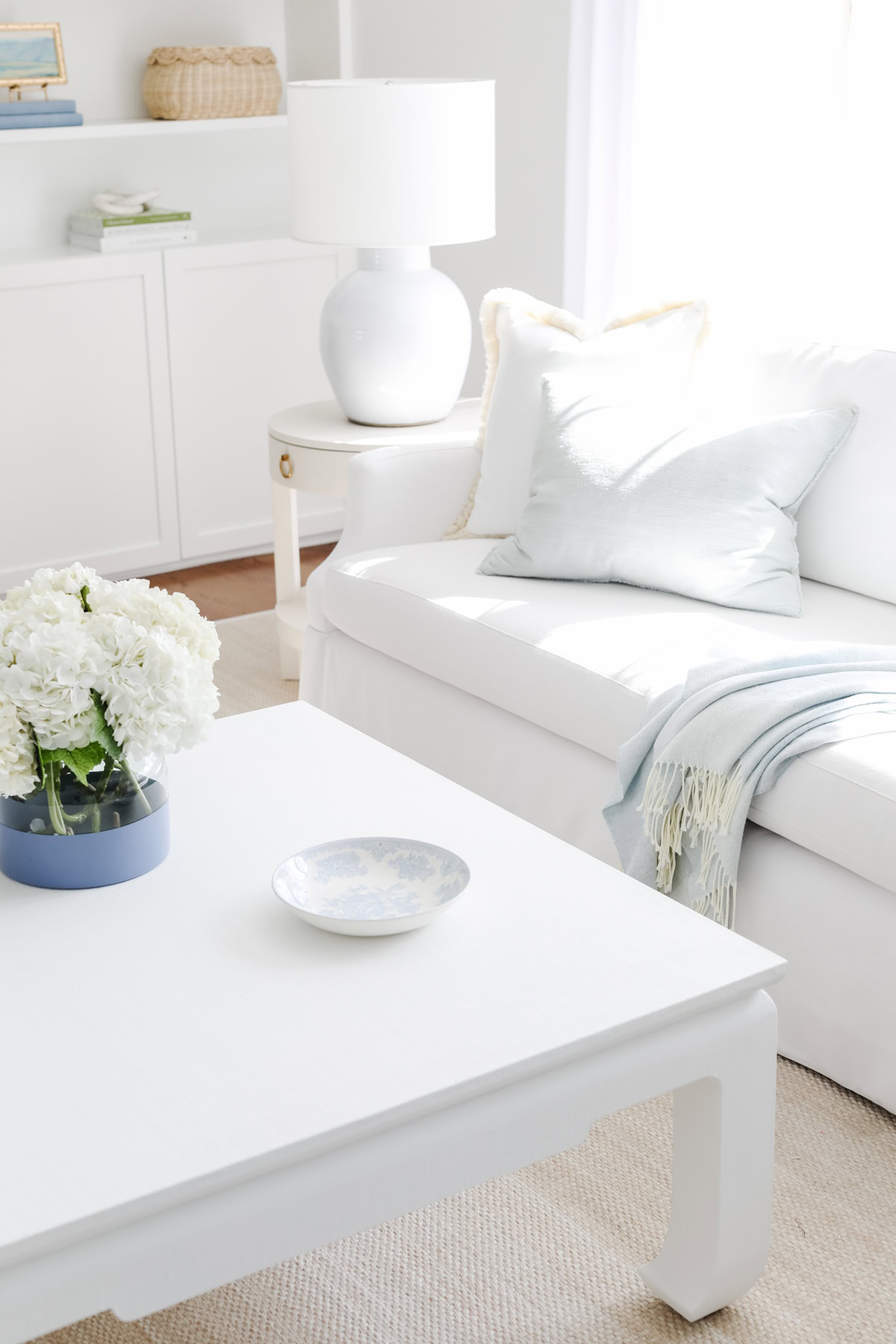 A white living room with a white sofa, light blue pillow, throw blanket, and a coffee table with flowers in a vase—perfect inspiration if you're learning how to measure furniture for the ideal fit in your own space. A lamp and basket complete the background.