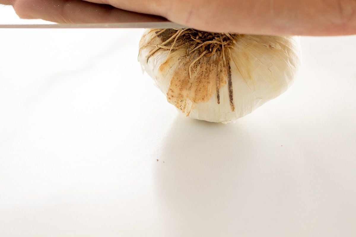 A hand pressing a large knife on top of a bulb of garlic