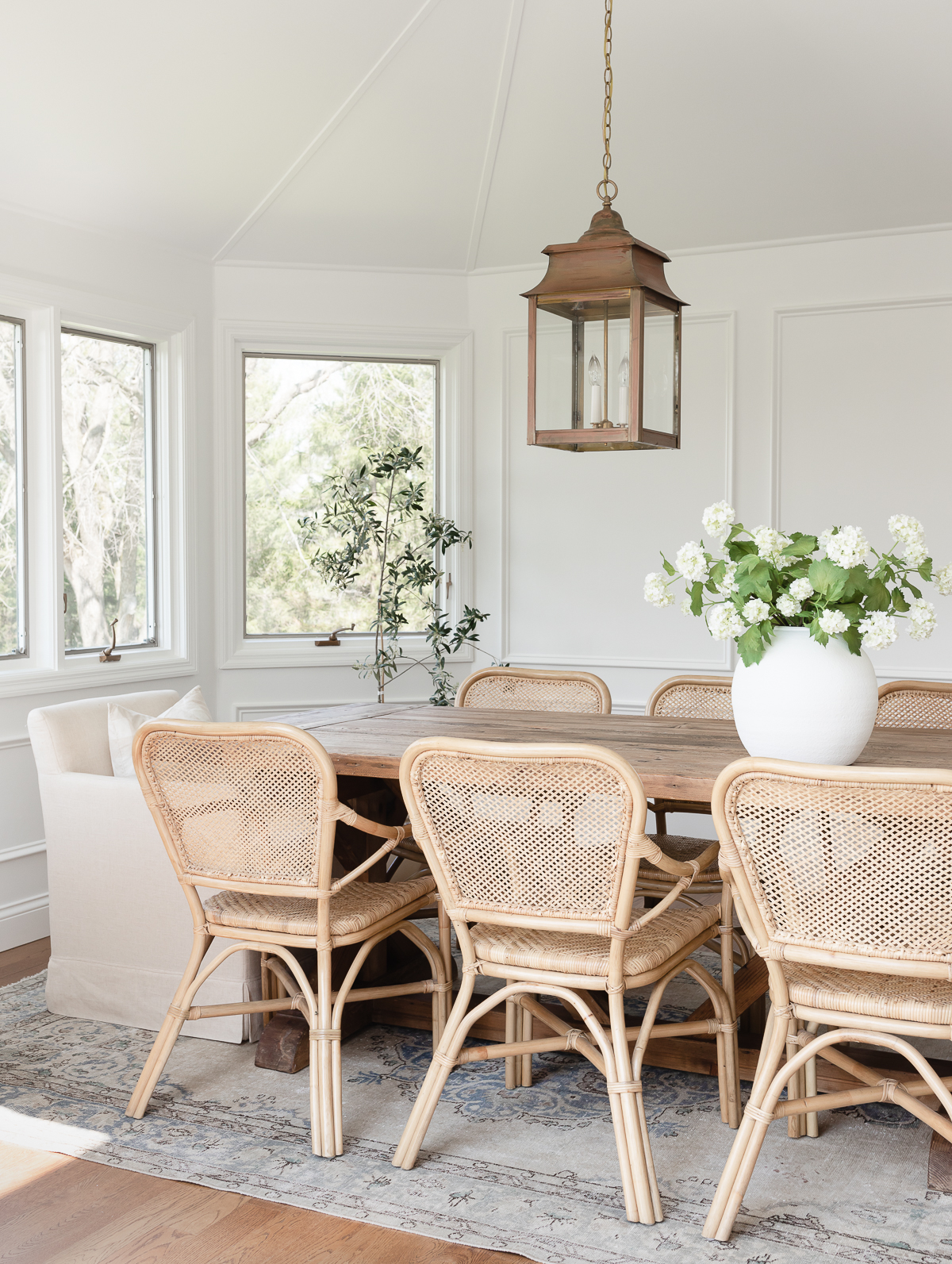A white dining room with a wood table, rattan chairs and picture frame moulding on the walls.