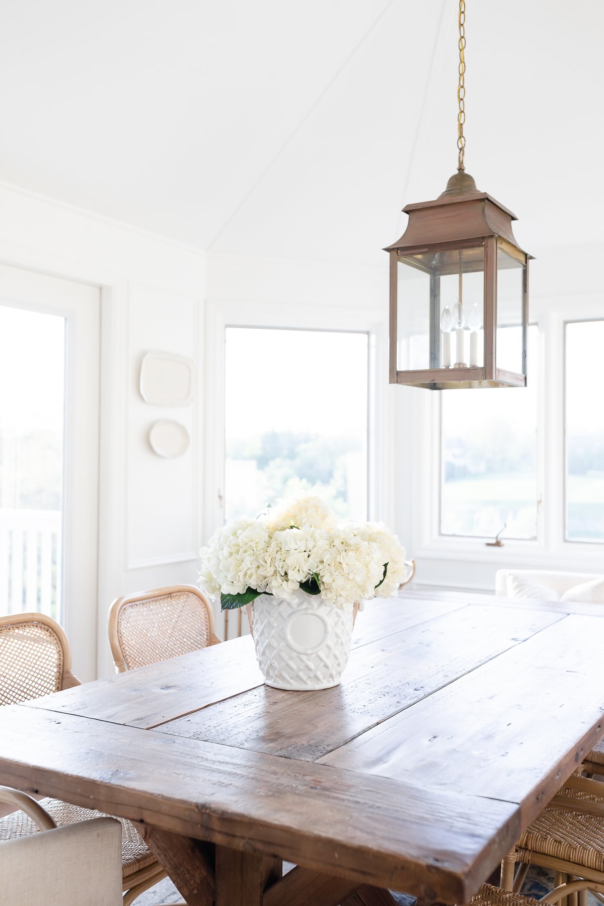 A white dining room with a wood table, rattan chairs and picture frame moulding on the walls.