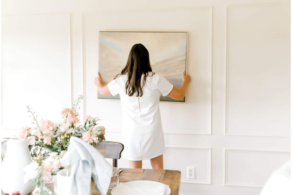 A woman hanging a painting in a white room with picture frame molding on walls